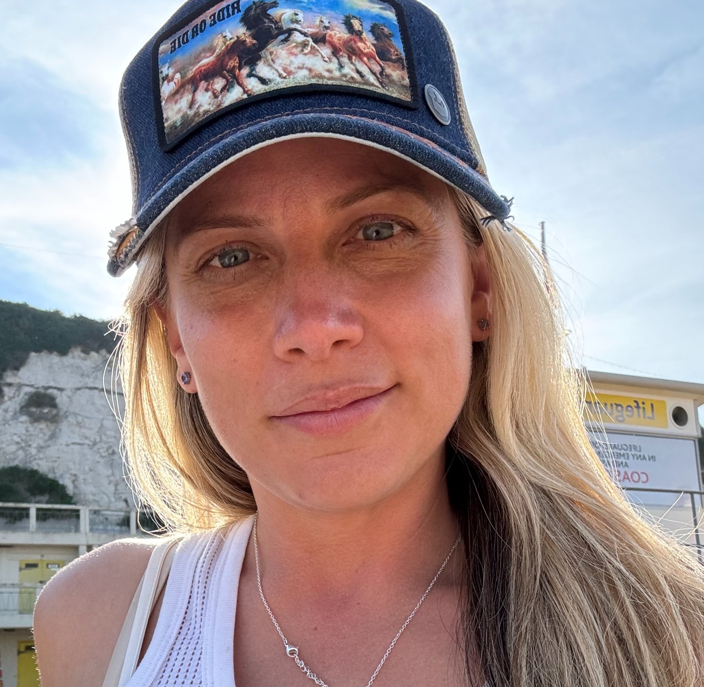 Tamsin Jones - with long blonde hair wearing a dark denim baseball cap with a patch of running horses and a sleeveless white top. She is outdoors on a sunny day, with white chalk cliffs, a yellow beachside building, and a blue sky in the background.